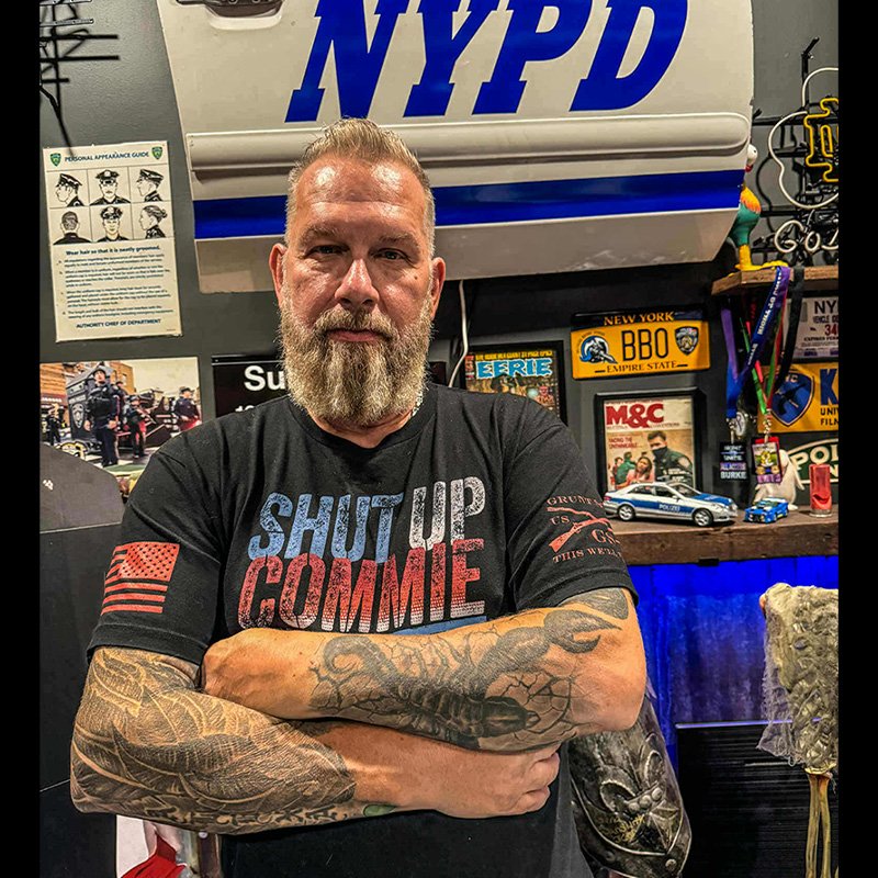 mike standing with his arms folded in front of a door from a NYPD car hanging on the wall behind him.